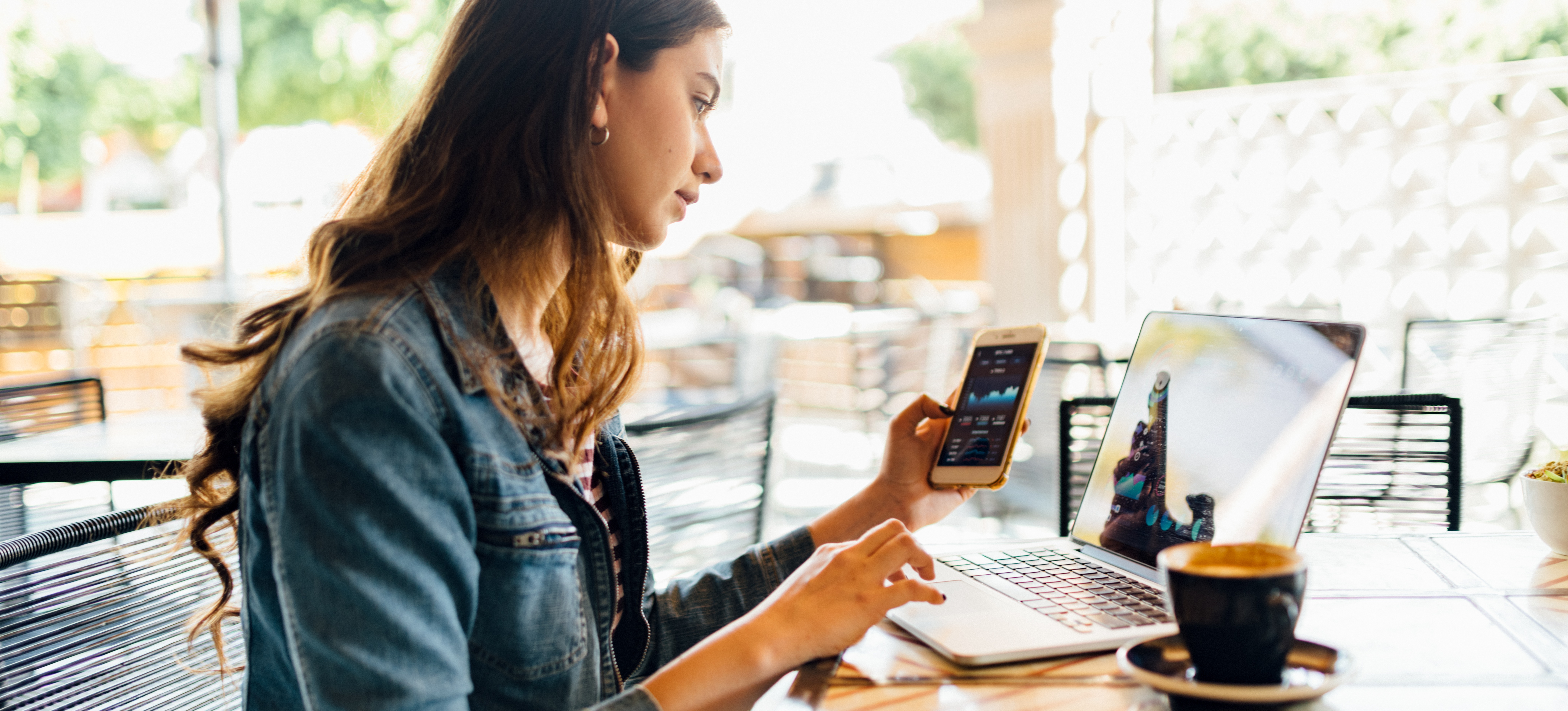[Featured Image]: Millennial student studying on their laptop and phone for their blockchain certification in an outdoor coffee shop.