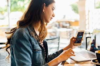 [Featured Image]: Millennial woman studying on her laptop and phone for her blockchain certification in an outdoor coffee shop.