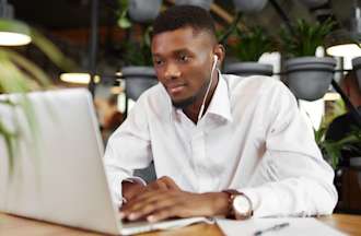 [Featured image] A social media manager in a white shirt wears earbuds and studies for a digital marketing certificate. They are seated at a table with a laptop and a potted plant. More potted plants are arranged behind them.