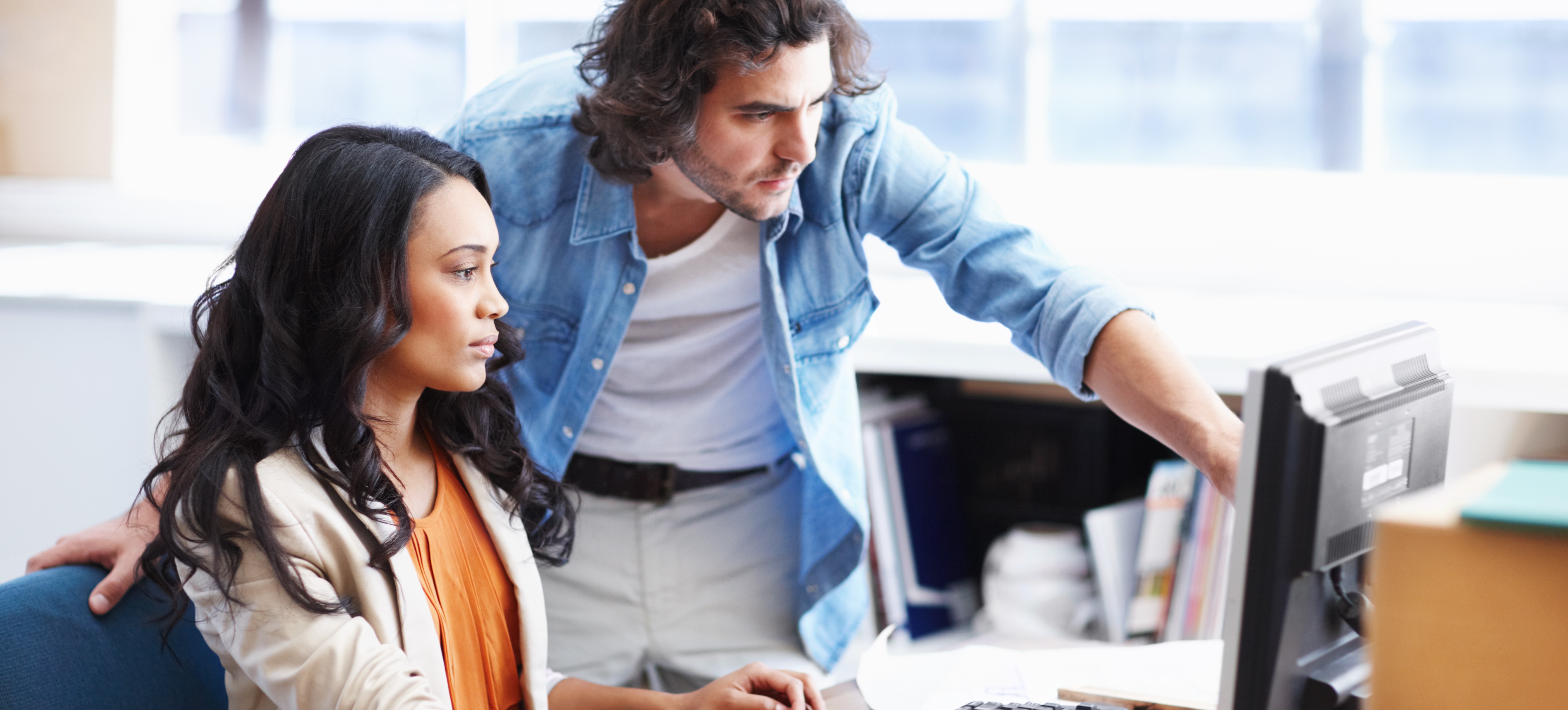 [Featured Image] Two colleagues look at open source software licensing on a computer screen in an office space near large windows.
