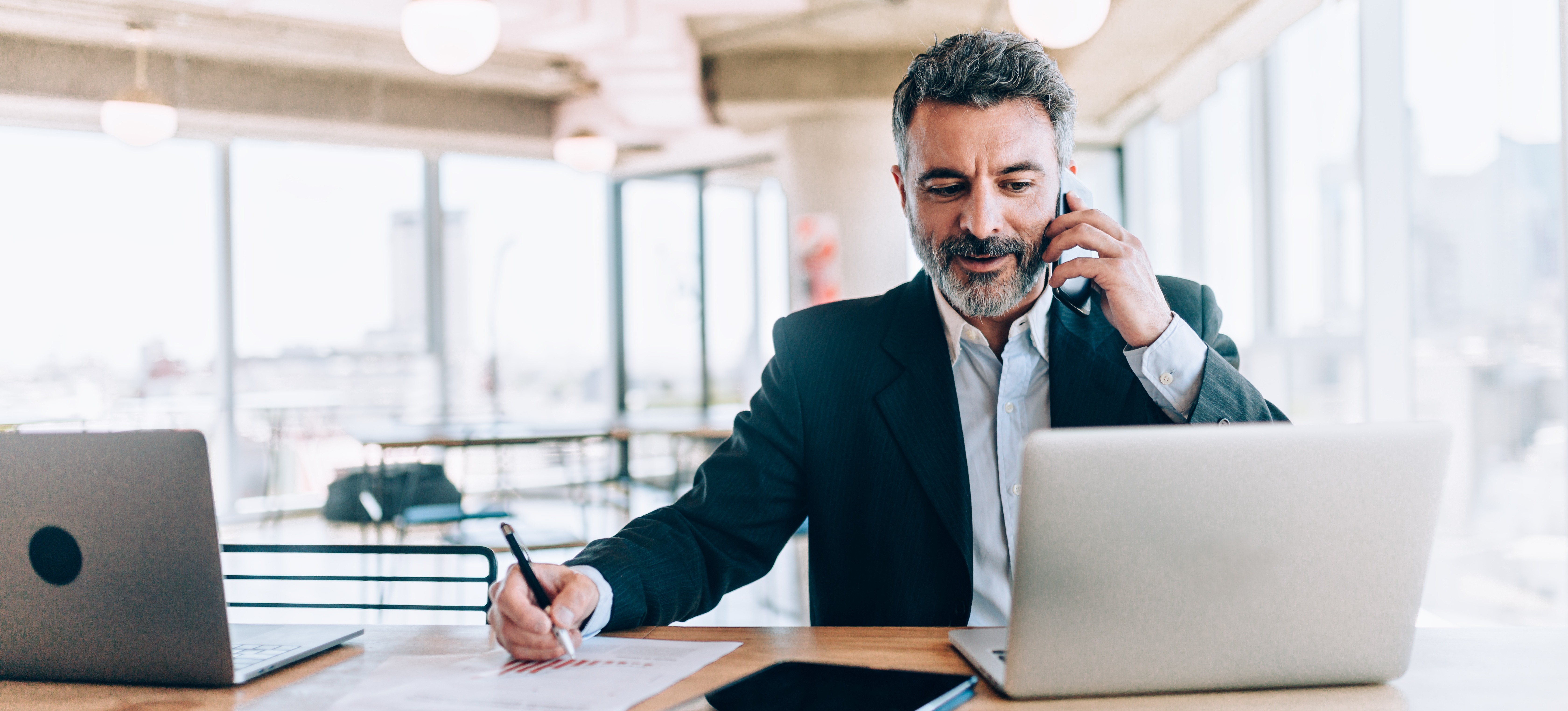 [Featured Image] A businessman working for a sales operation specialist salary sits at a desk in an open office, looking at his laptop while talking on his cell phone and writing on a piece of paper.
