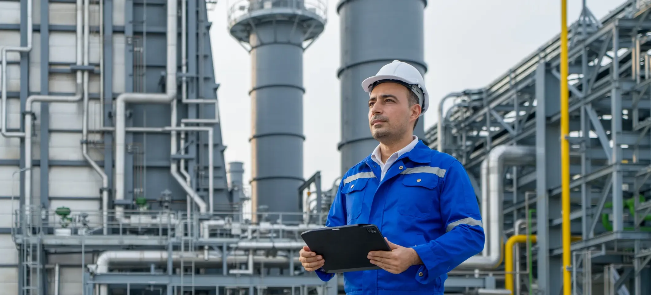 [Featured Image] An engineer wearing a hard hat who has experience with cybersecurity in the oil and gas industry holds a tablet while standing in front of a petroleum oil refinery.
