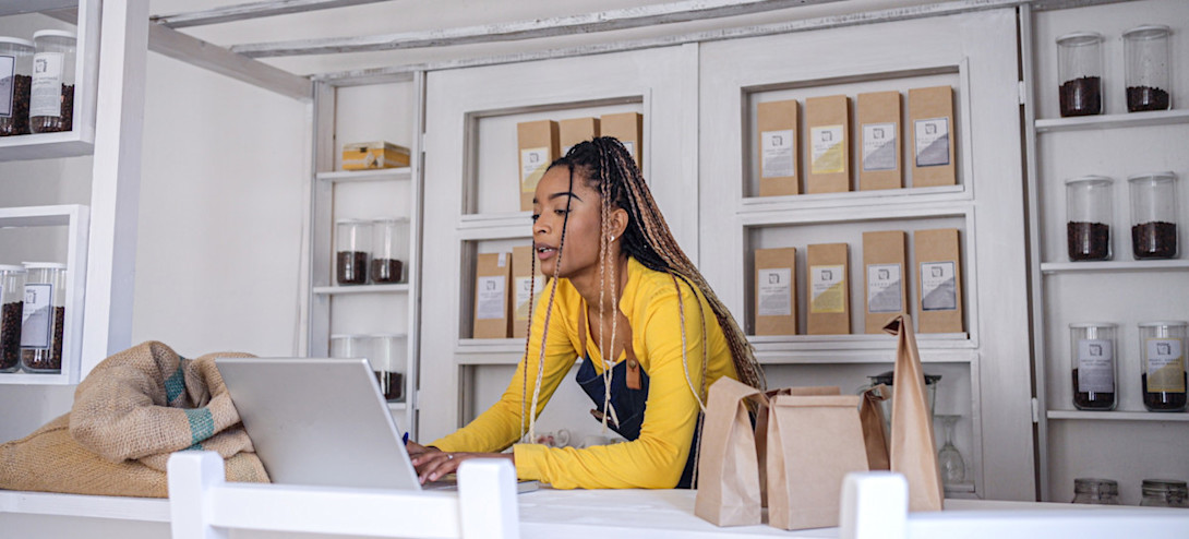 [Featured Image] An eco-friendly coffee shop owner sets up a Google search campaign on their laptop as they stand at the counter in their shop.
