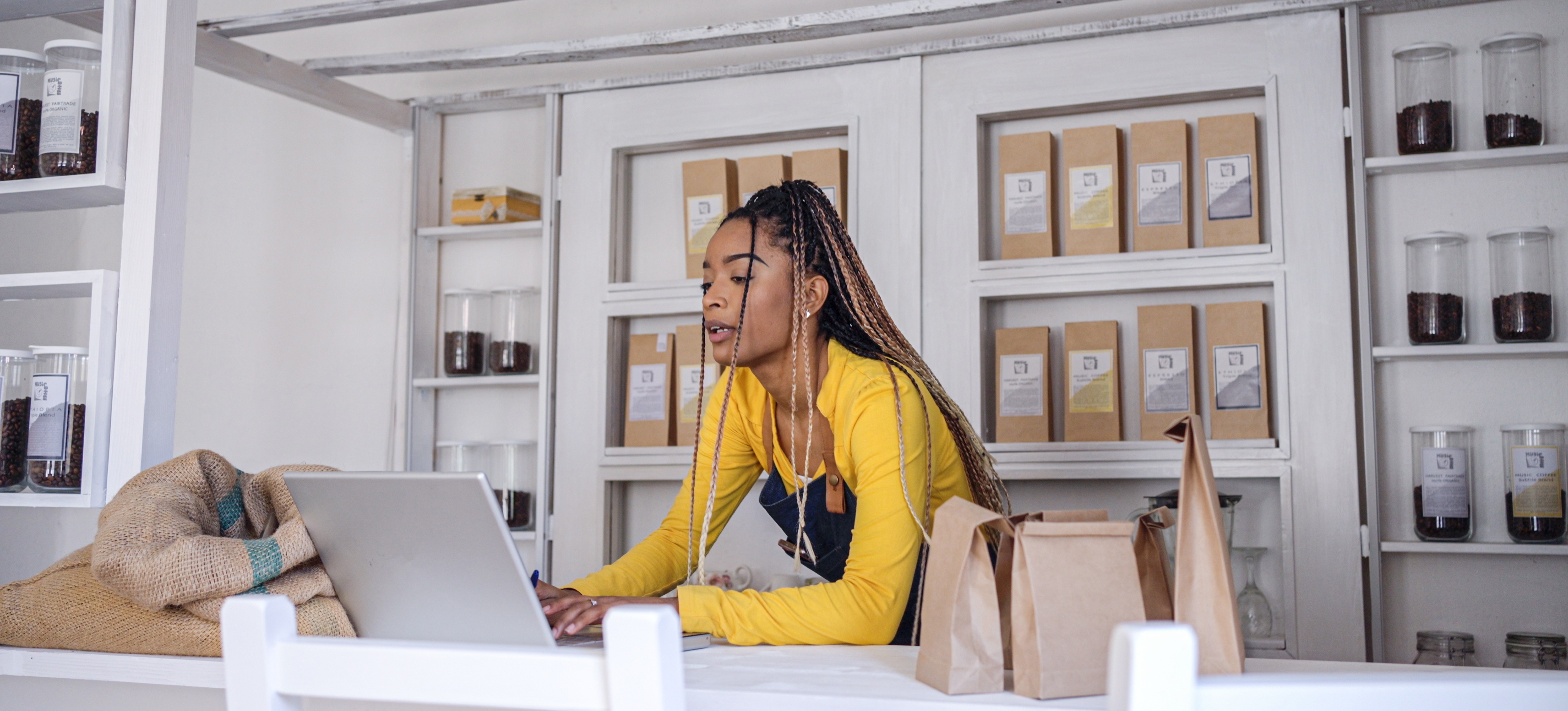 [Featured Image] An eco-friendly coffee shop owner sets up a Google search campaign on their laptop as they stand at the counter in their shop.
