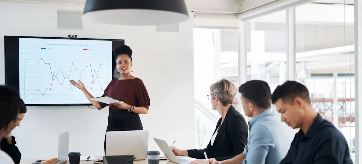 [Featured Image] A brand manager points to a chart as they discuss brand marketing with five people at a conference table. 