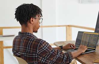 [Featured Image] A python developer looks at code on his laptop and desktop computer in his home office.