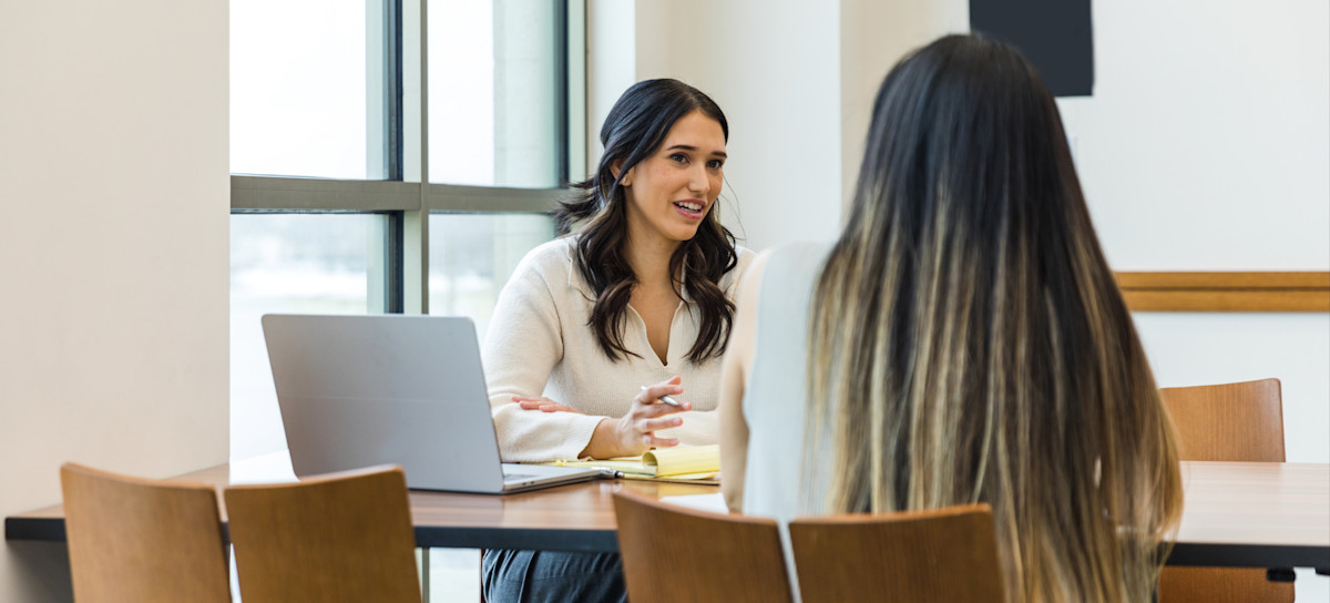 [Featured Image] A person working for a career counselor salary talks to a younger person at a table, with a laptop and notebook between them.
