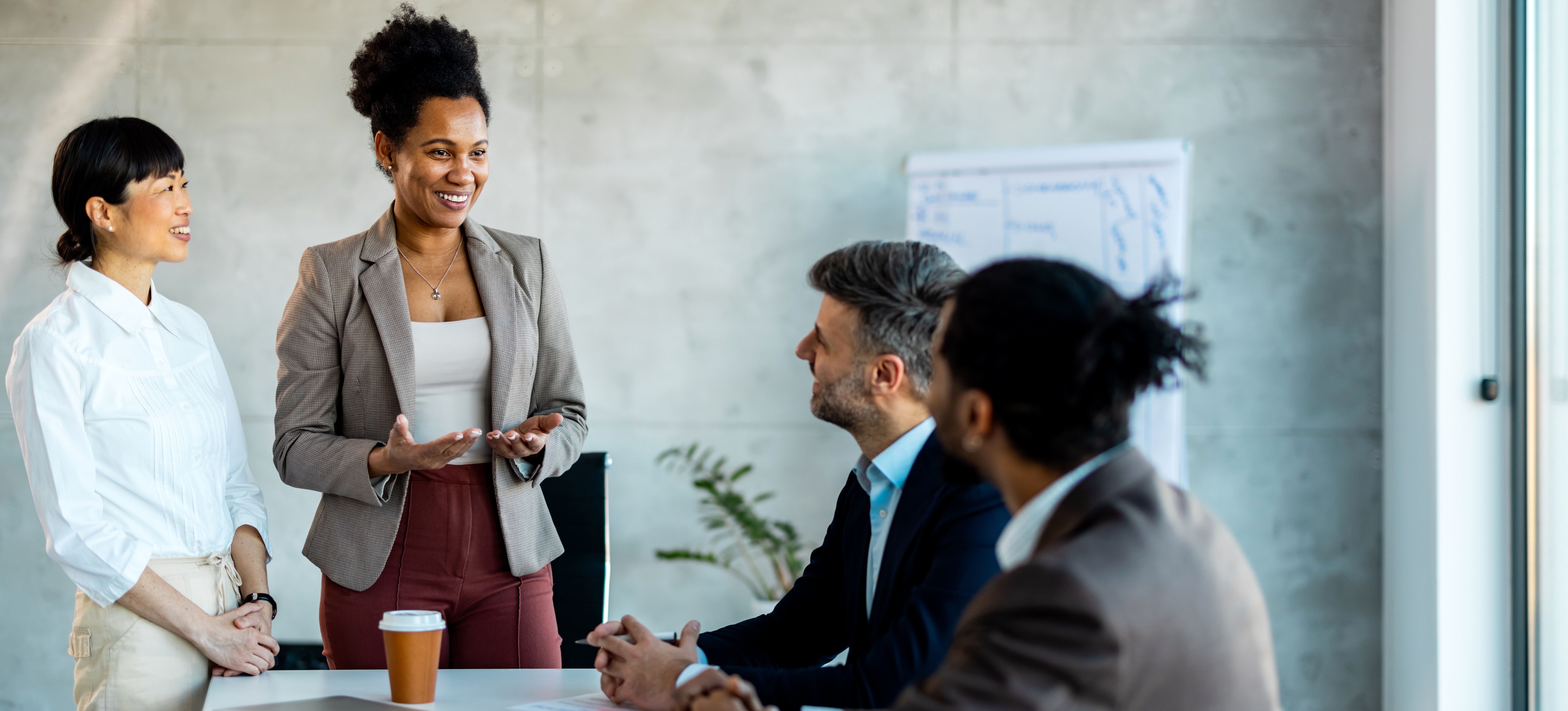 [Featured Image] A smiling human resources manager uses HR leadership skills as they stand in front of their colleagues sitting at a boardroom table and explain team concepts.