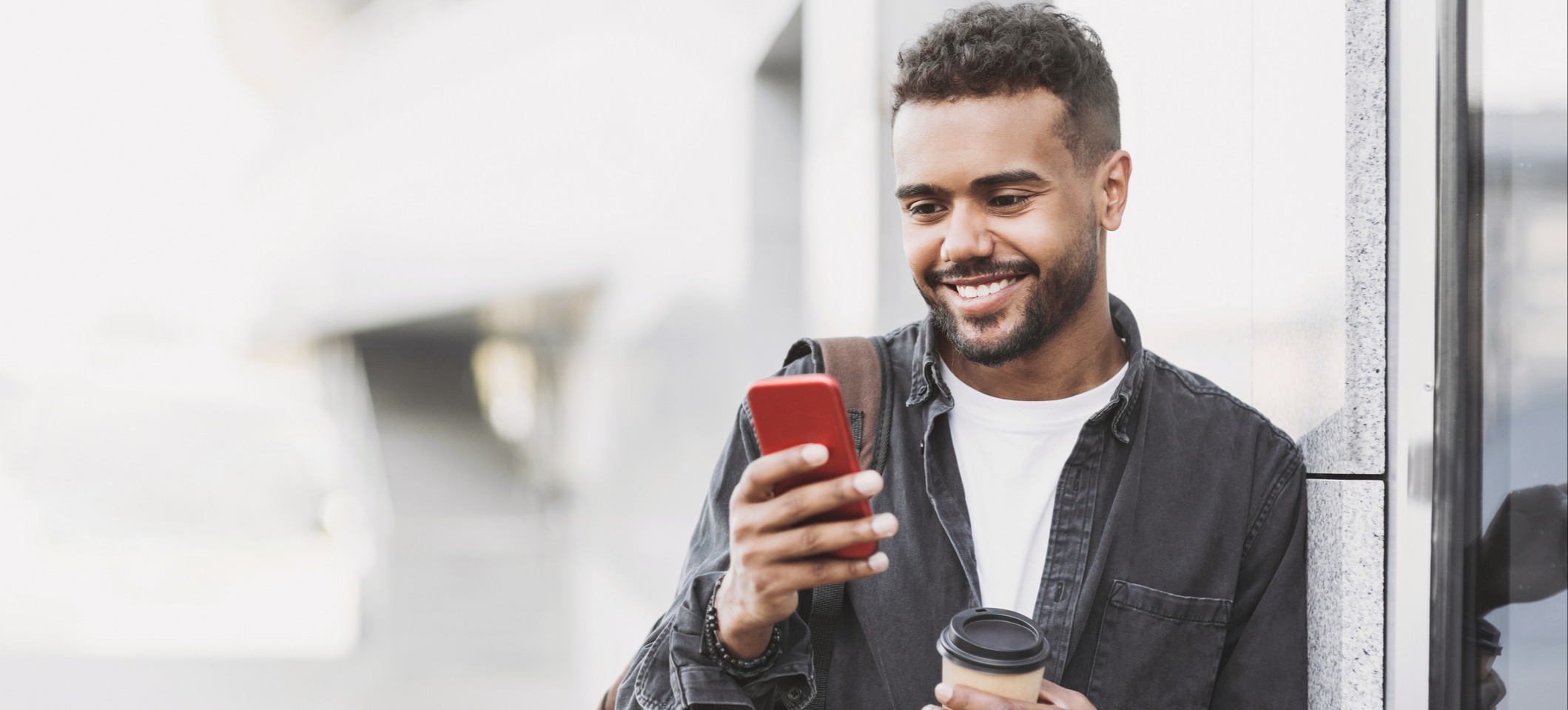 [Featured Image] A person stands outside a building holding a to-go coffee and listening to a podcast on his phone.