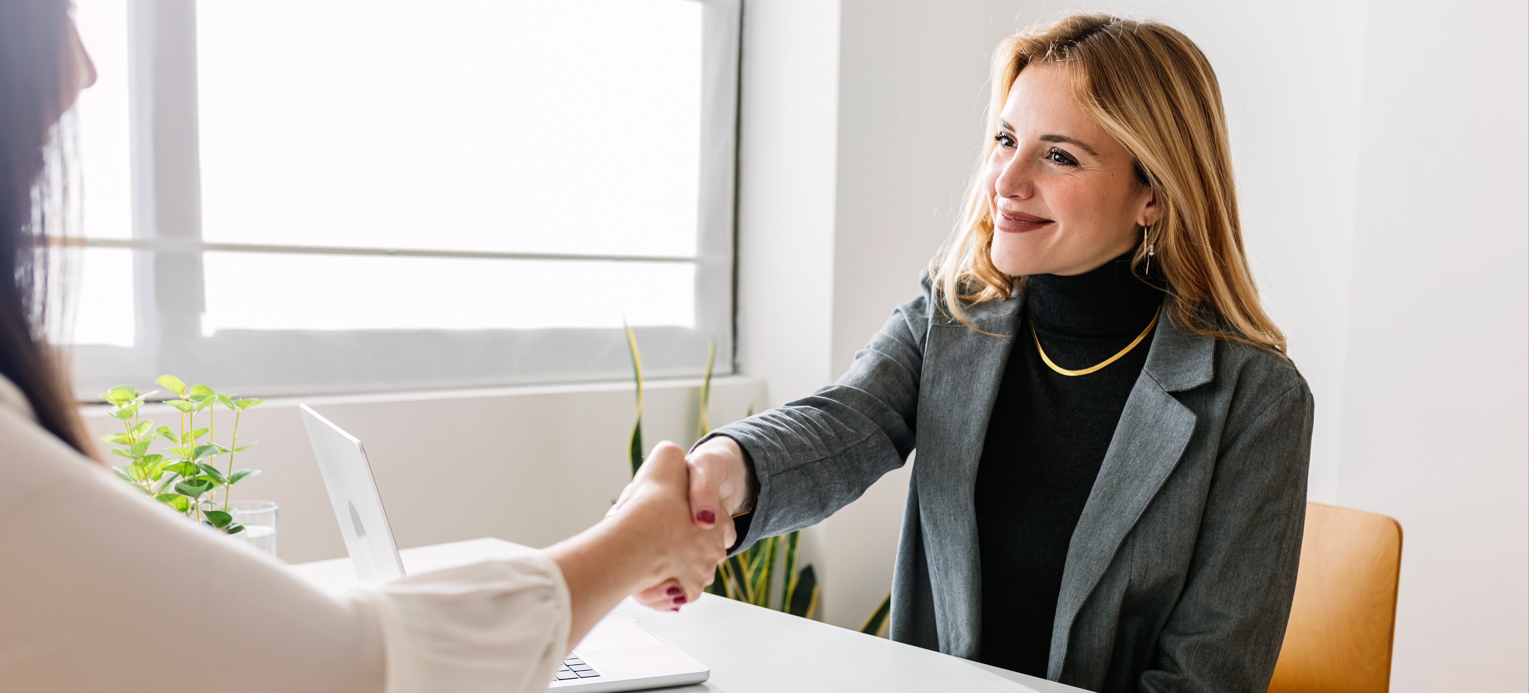 [Featured Image] A woman wearing a gray blazer and a black turtleneck is shaking hands with her job interviewer across a desk and wondering how long interviews last.
