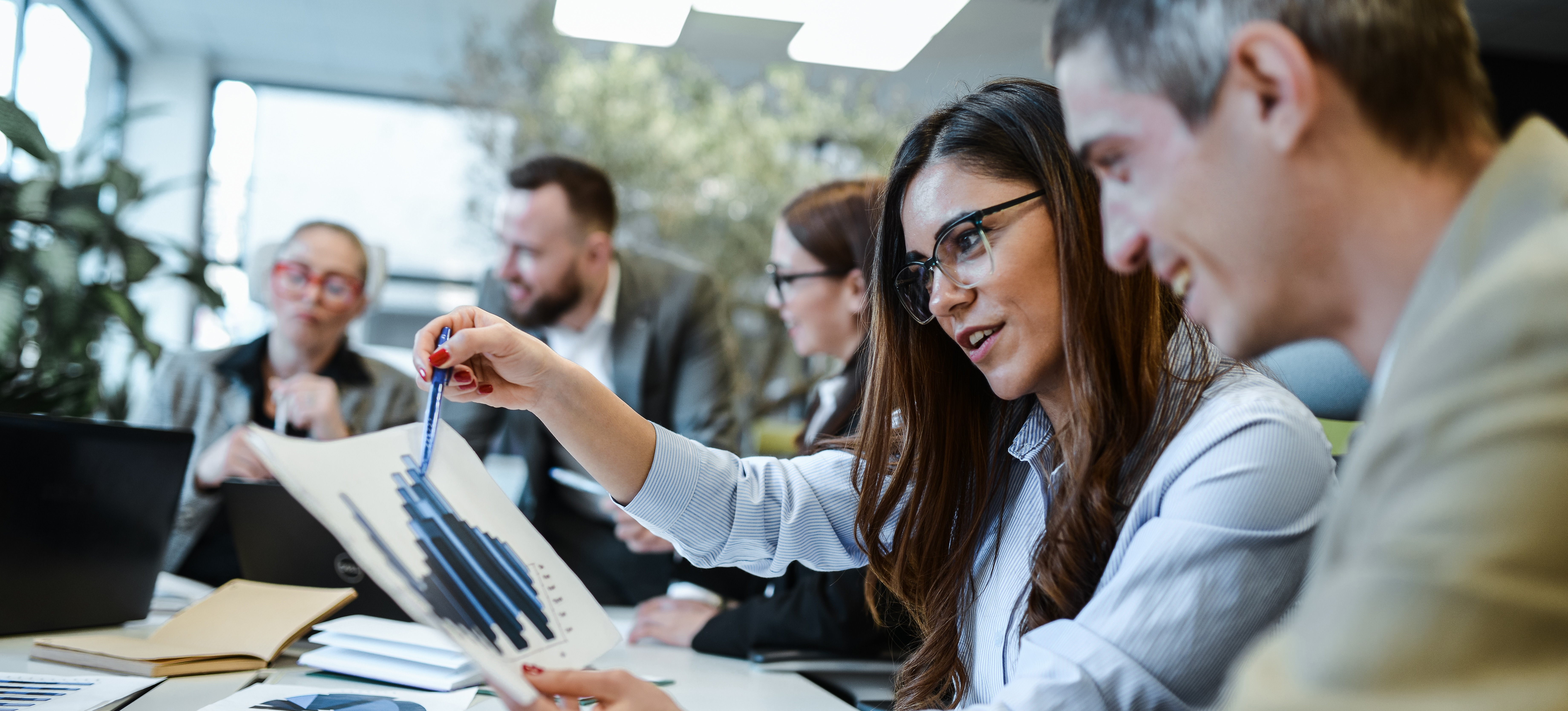 [Featured Image] A project manager sits with a colleague and explains a graph on a report while other team members collaborate in the background.
