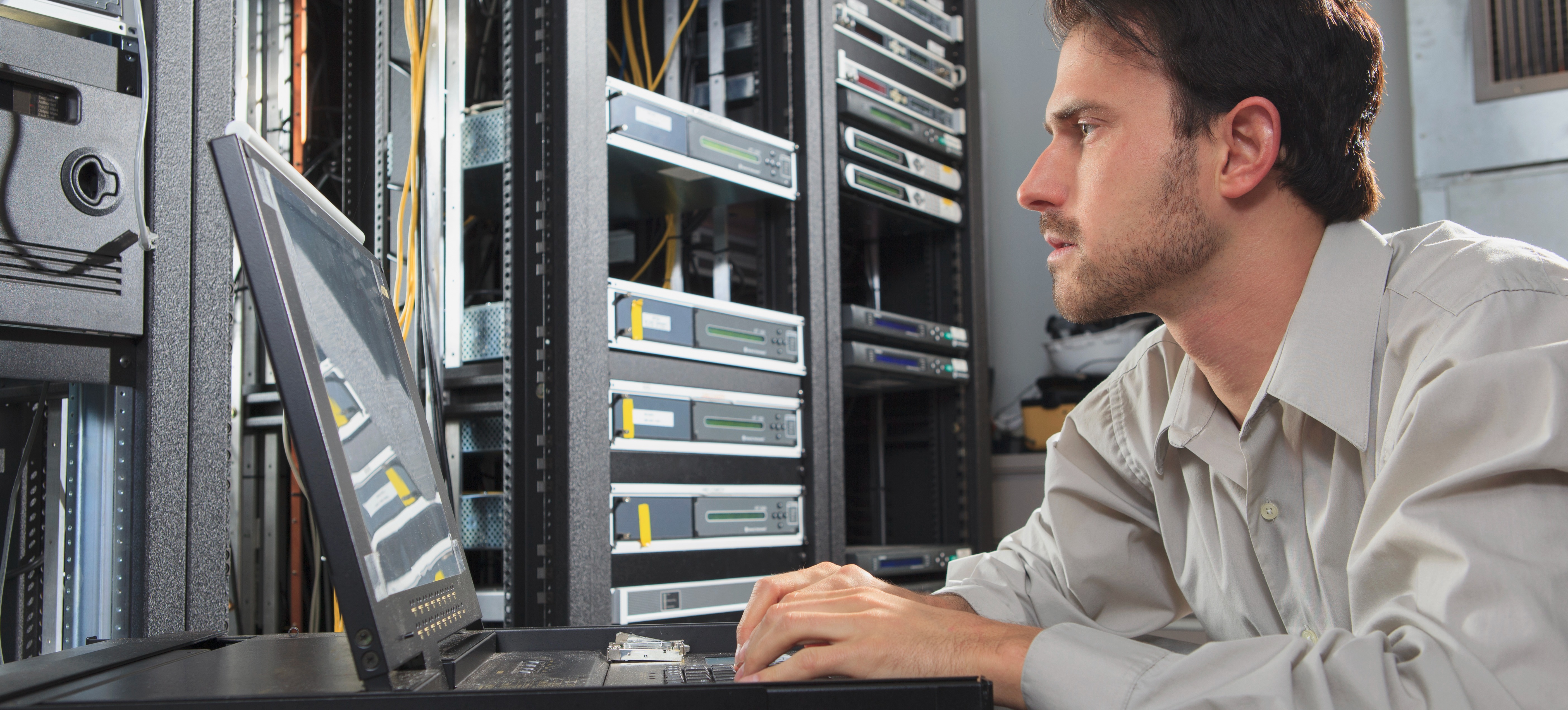 [Featured Image] A network security engineer looking at a monitor in a cable server room and programming configurations.