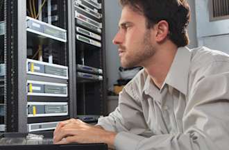 [Featured Image] A male network security engineer looking at a monitor in a cable server room and programming configurations.