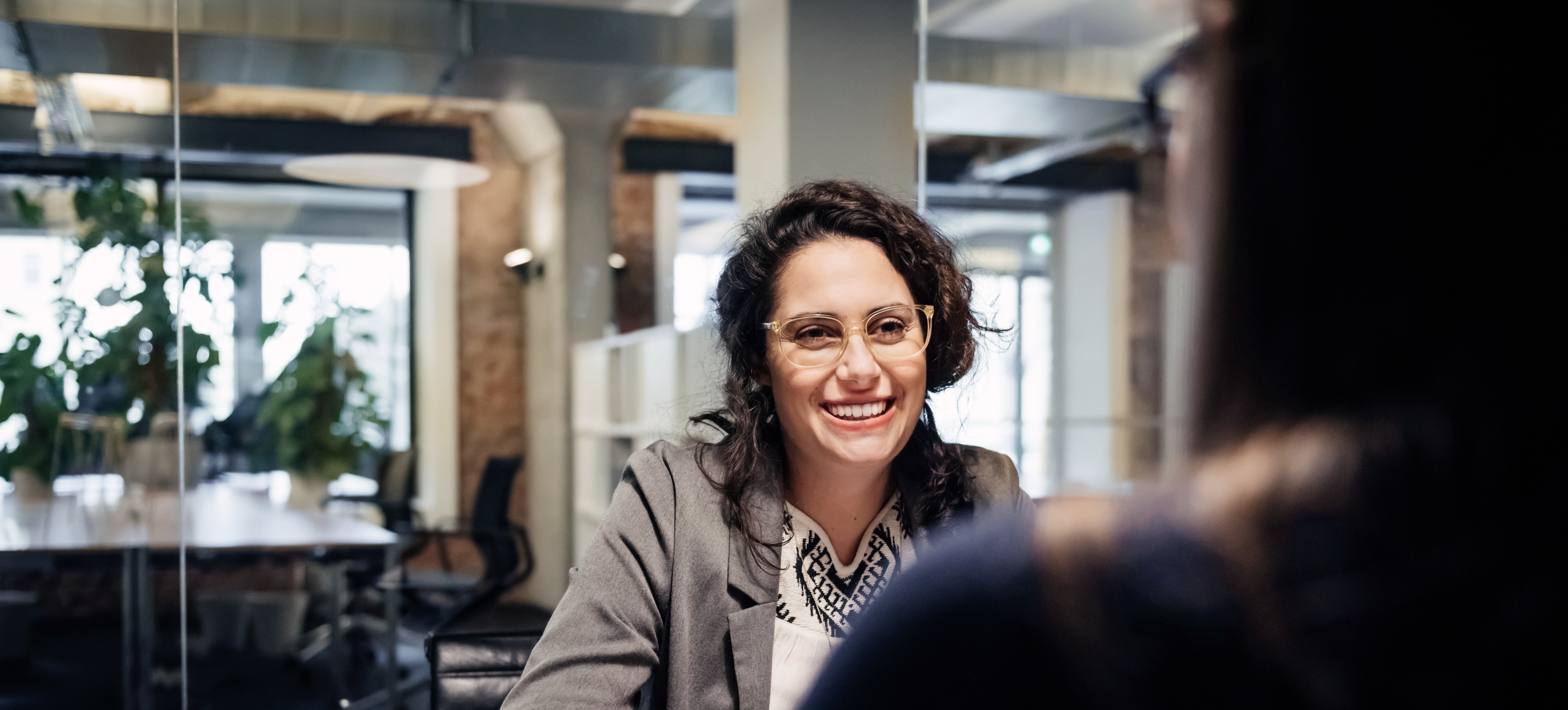 [Featured Image] A person wearing glasses, a grey jacket, and a white blouse sits across a desk talking to a person wearing a blue blouse and glasses.