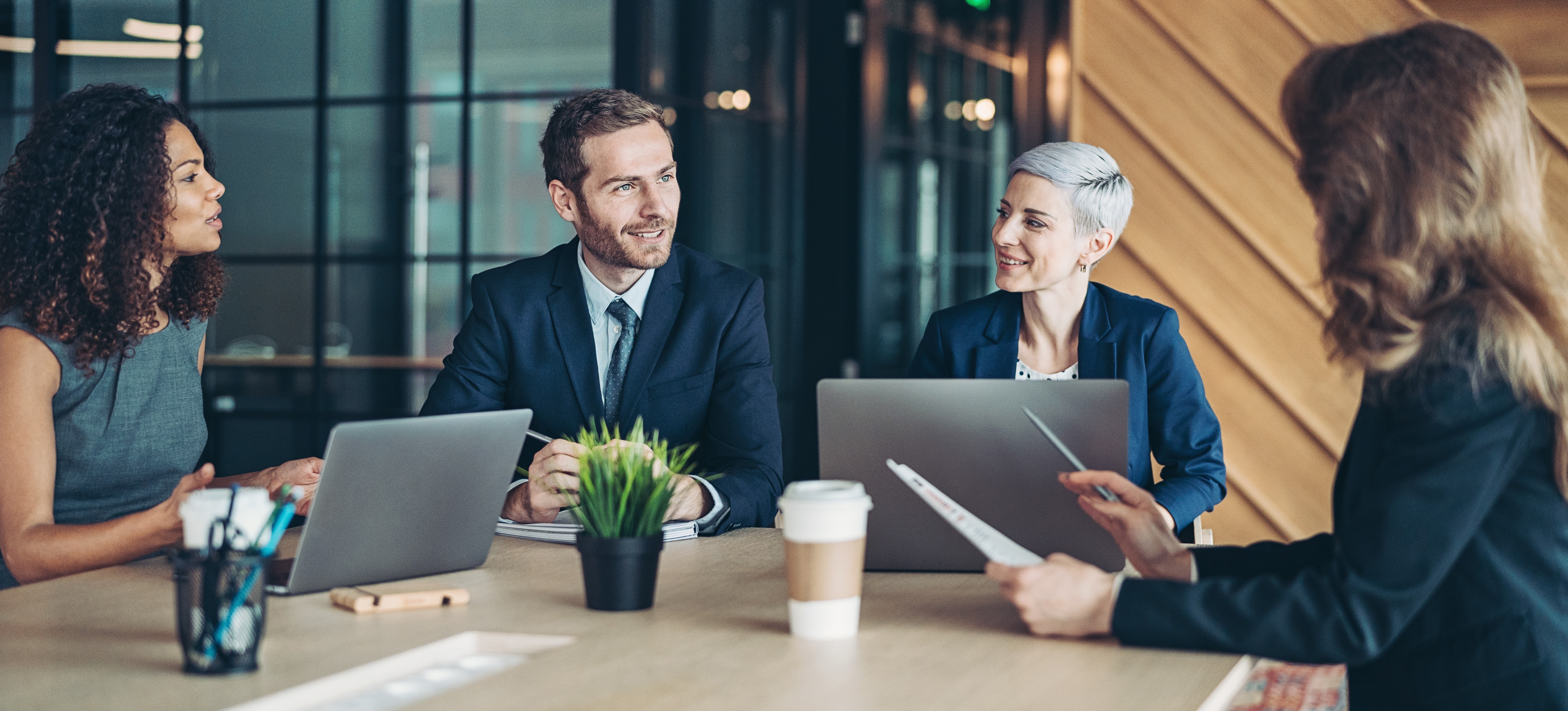 [Featured Image] Four professionals who have earned finance certifications meet in an office around a table using their laptops.

