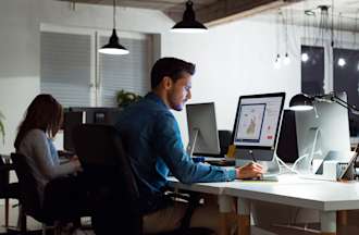 [Featured image] Two people sit next to each other at a shared desk and work on separate computers in a dimly lit open office space.