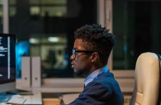 [Featured Image]: A man wearing a blue sweater and glasses, is working in front of two computer screens. He is sitting on a creamy white chair. 