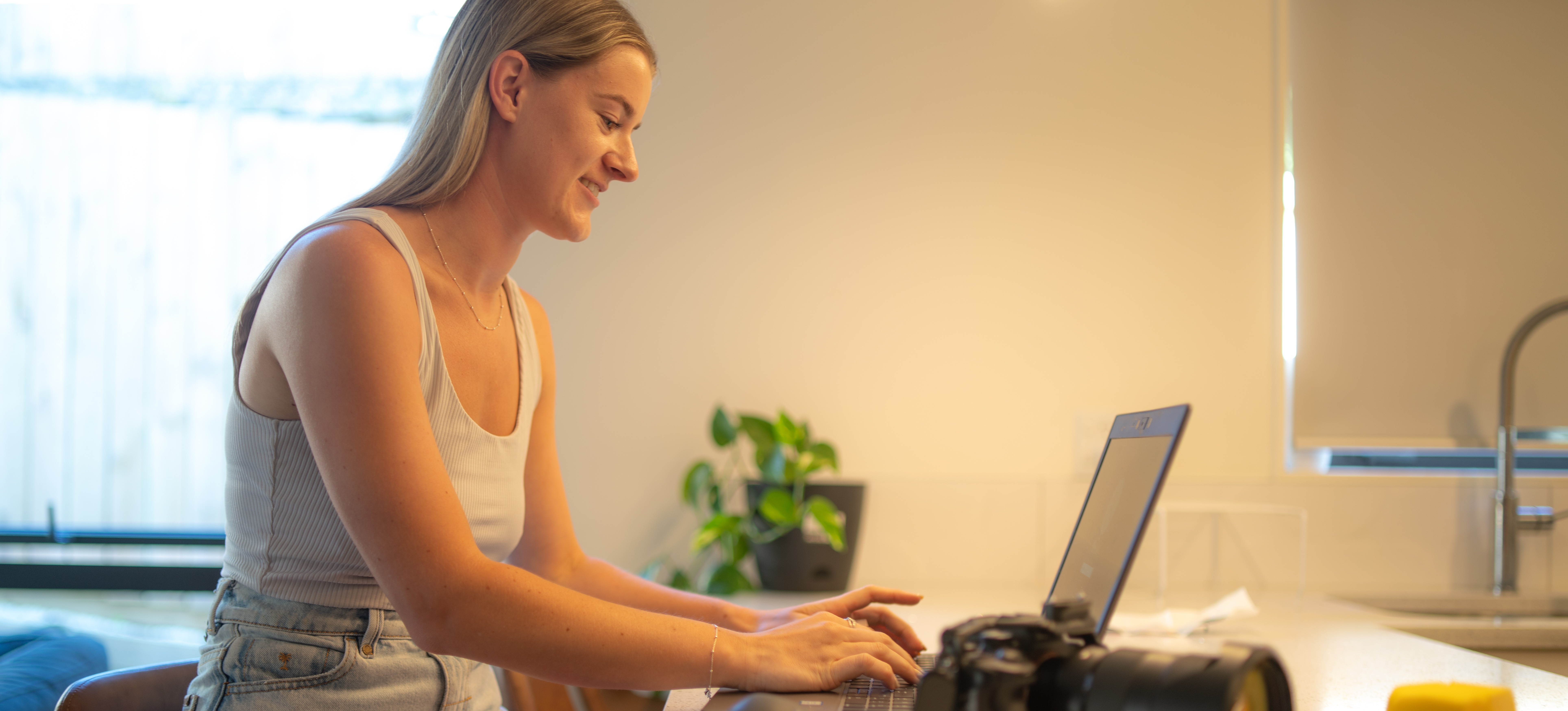 [Hauptbild] Eine digitale Schöpferin in einem grauen Tanktop arbeitet an ihrem Schreibtisch zu Hause mit ihrem Laptop und ihrer Digitalkamera an einem Projekt.