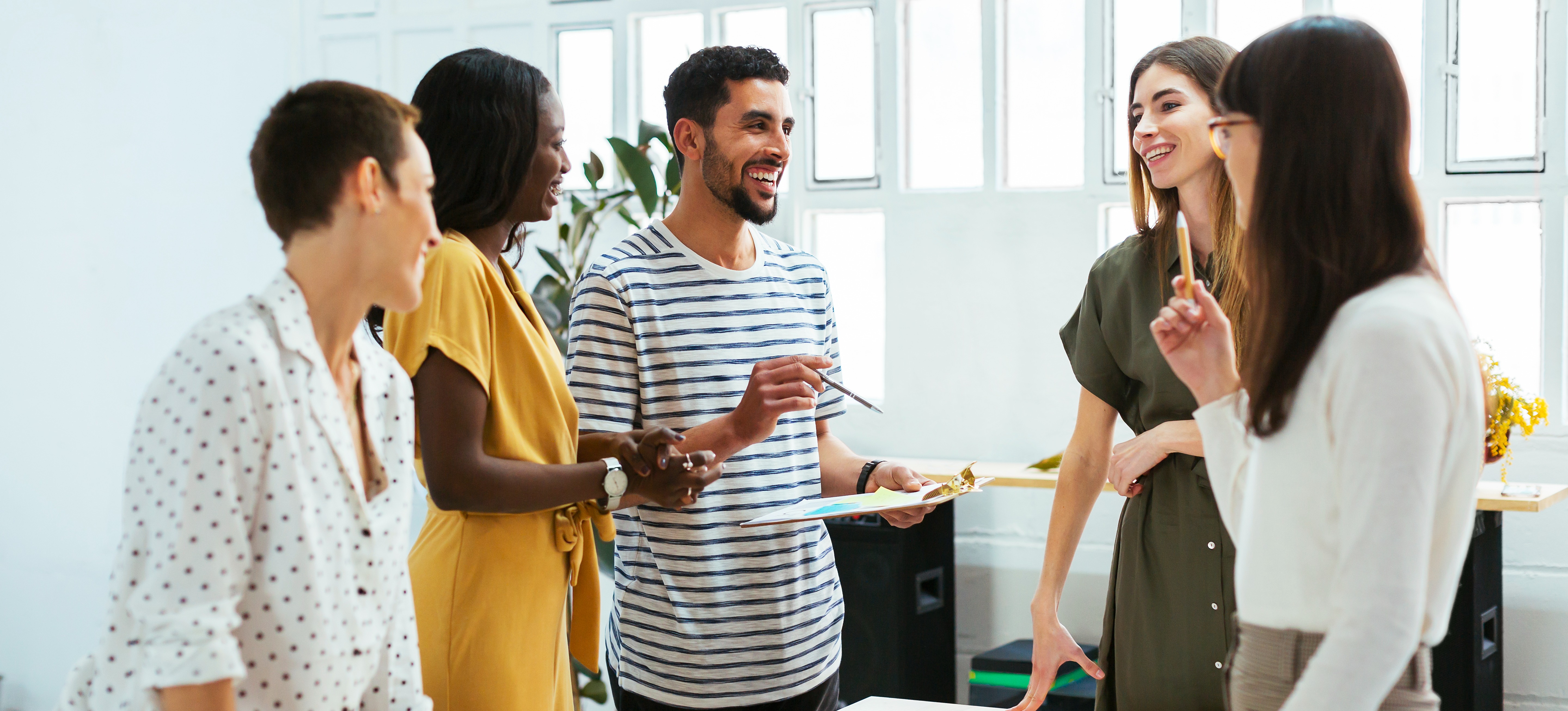 [Featured Image] A smiling social media coordinator stands with his team in a conference room discussing the company's social media strategy.