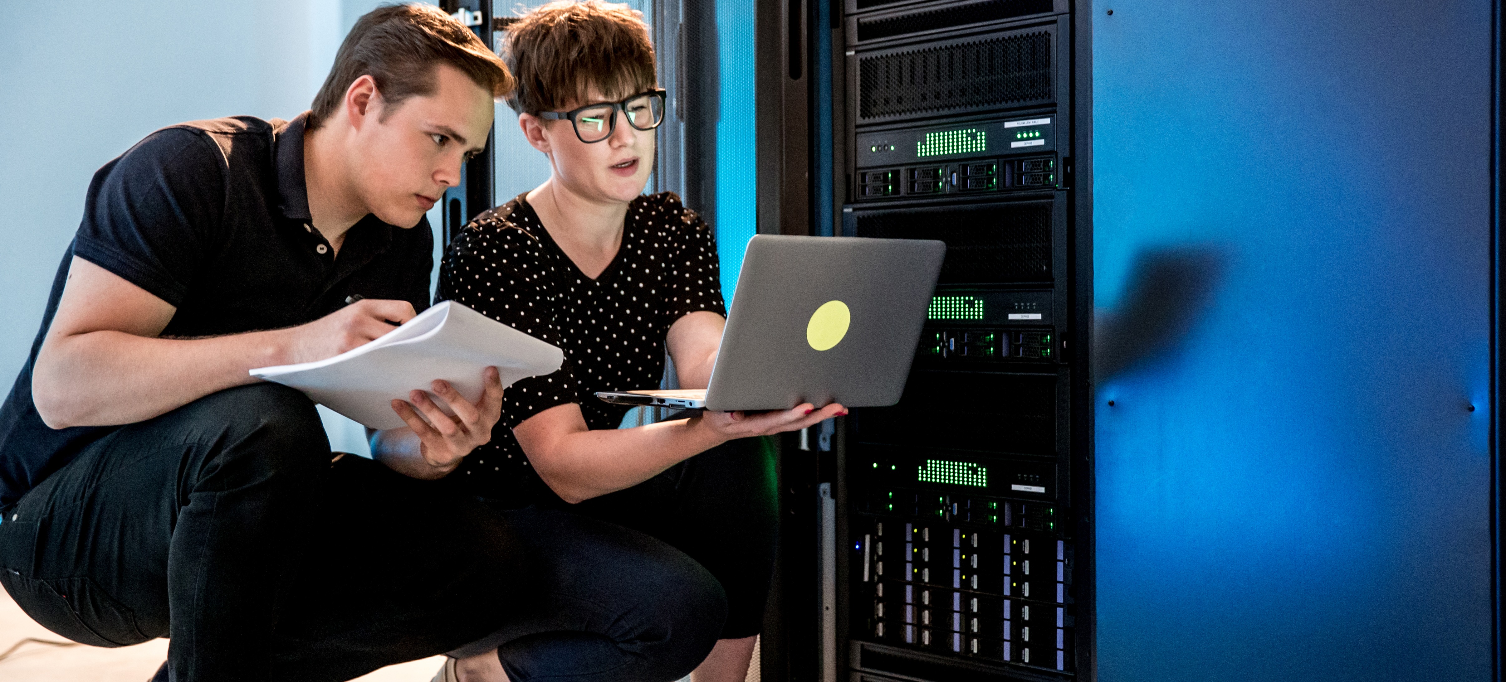 [Hauptbild] Zwei Sicherheitstechniker arbeiten in einem Serverraum mit einem Laptop und Dokumenten.
