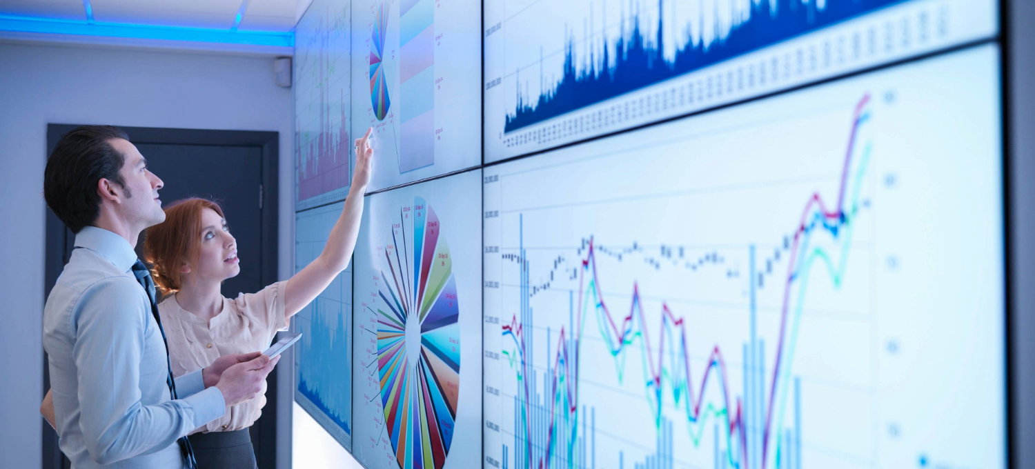 [Featured image] Two colleagues stand in front of several graphs and analyze the data for their supply chain. 