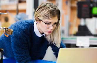 [Featured image] A woman wearing glasses stands over a robotics work table staring at her laptop.