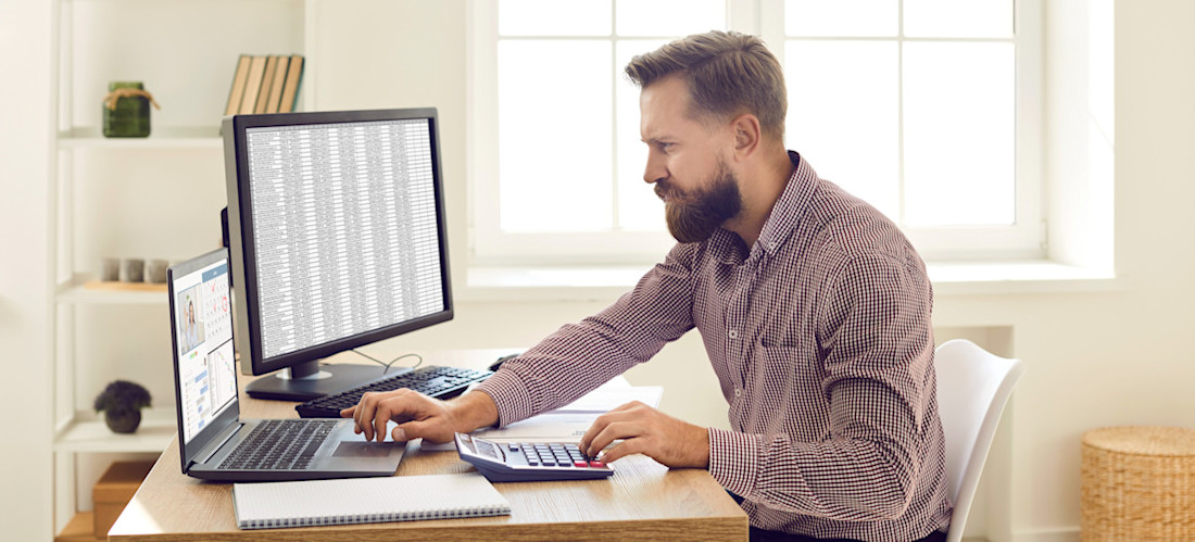 [Featured Image] A person in their home office is using a Google Sheets solver application while looking at a computer monitor and typing on a laptop and manual calculator.
