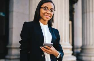 [Featured image] An MBA student stands outside a columned building holding her mobile phone and looking at the camera.