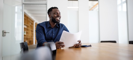 [Featured Image] A man in a blue button-up is sitting down in a conference room holding pieces of paper. 