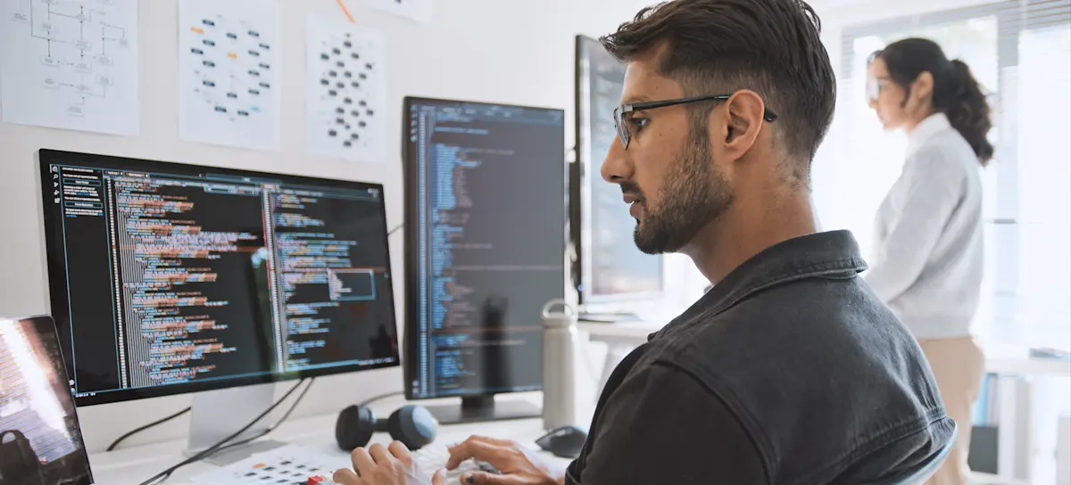 [Featured Image] A QA tester sits at several computer monitors displaying data while their colleague stands in the background and works at computer monitors.
