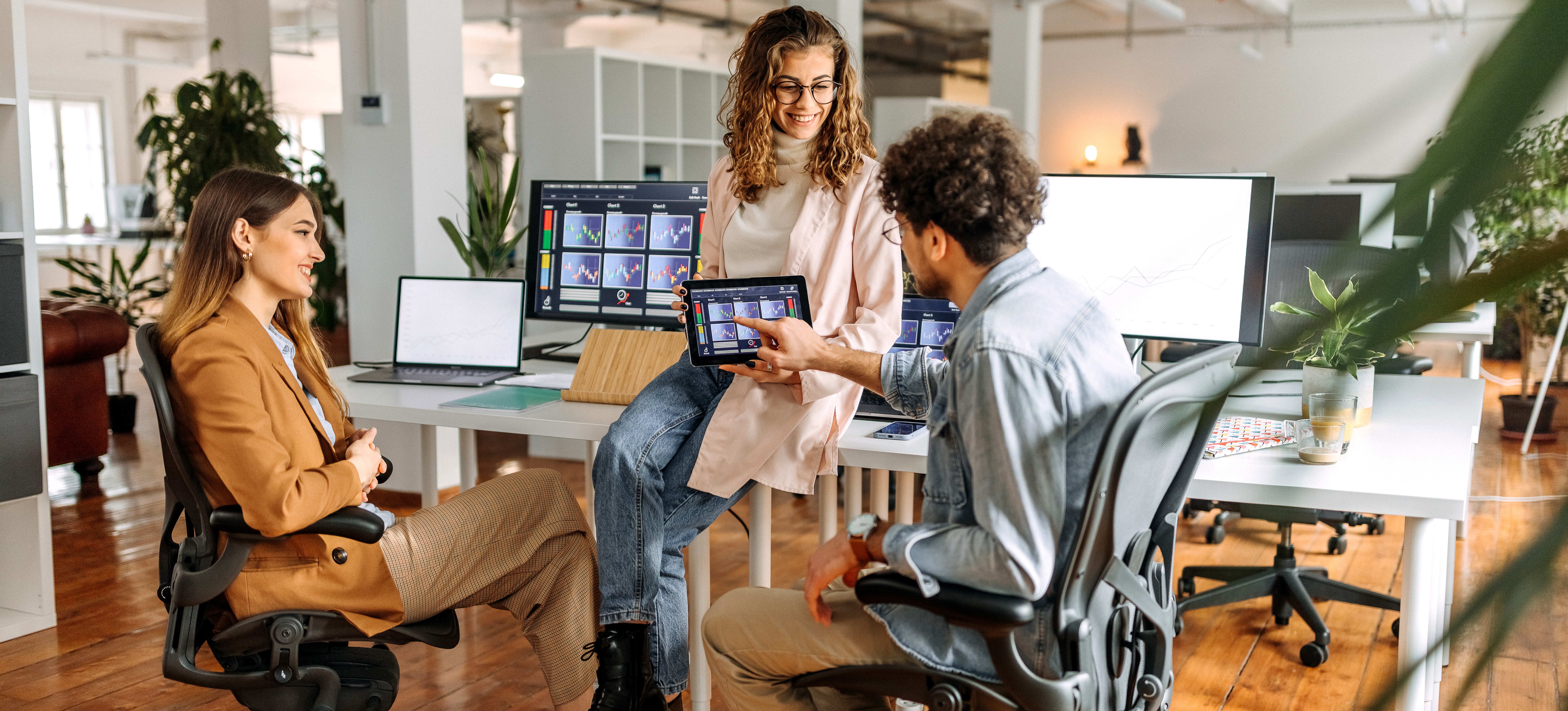 [Featured Image] A group of young coworkers sit in a circle at their desks as one shows the others virtual currency data on a tablet.
