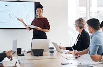 [Featured image] A women stands in front of a digital whiteboard and leads a marketing strategy meeting with several coworkers. 