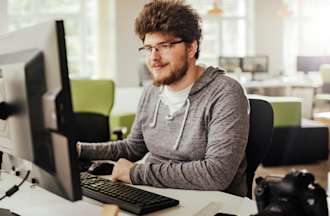 [Featured image] An AWS DevOps engineer in a gray hoodie and glasses sits at a desktop computer in a shared workspace.