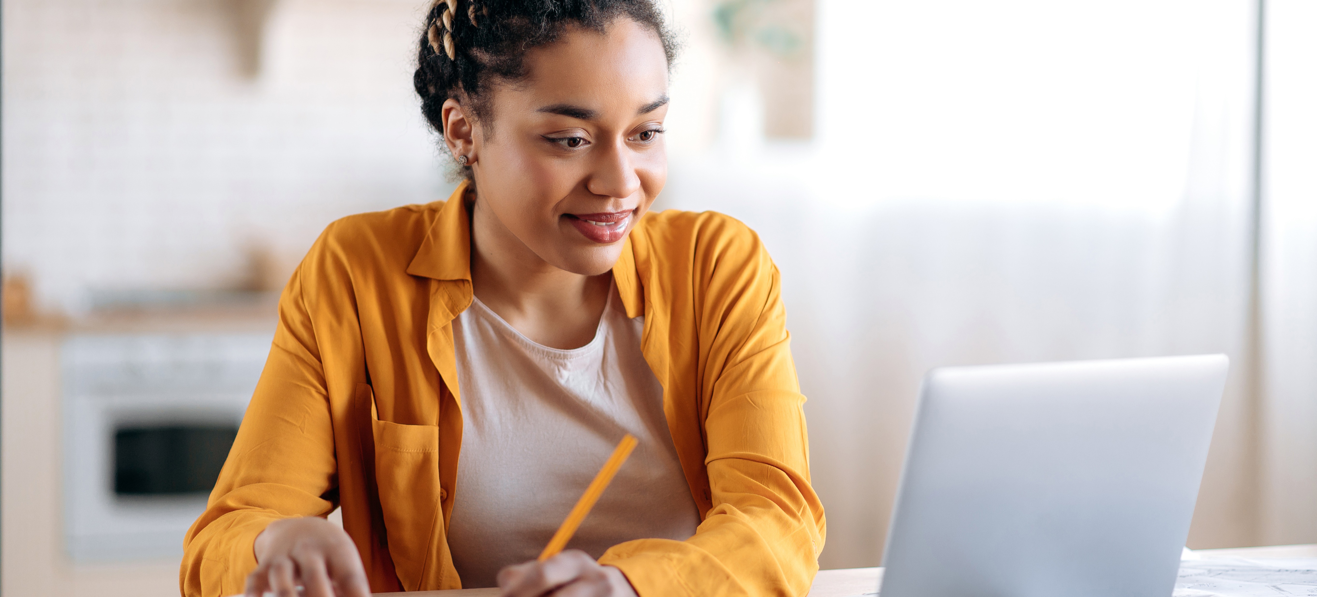 [Featured Image] A young student in a yellow shirt sits at a table, writing in a notebook and looking at a laptop, as she studies for the PSAT.
