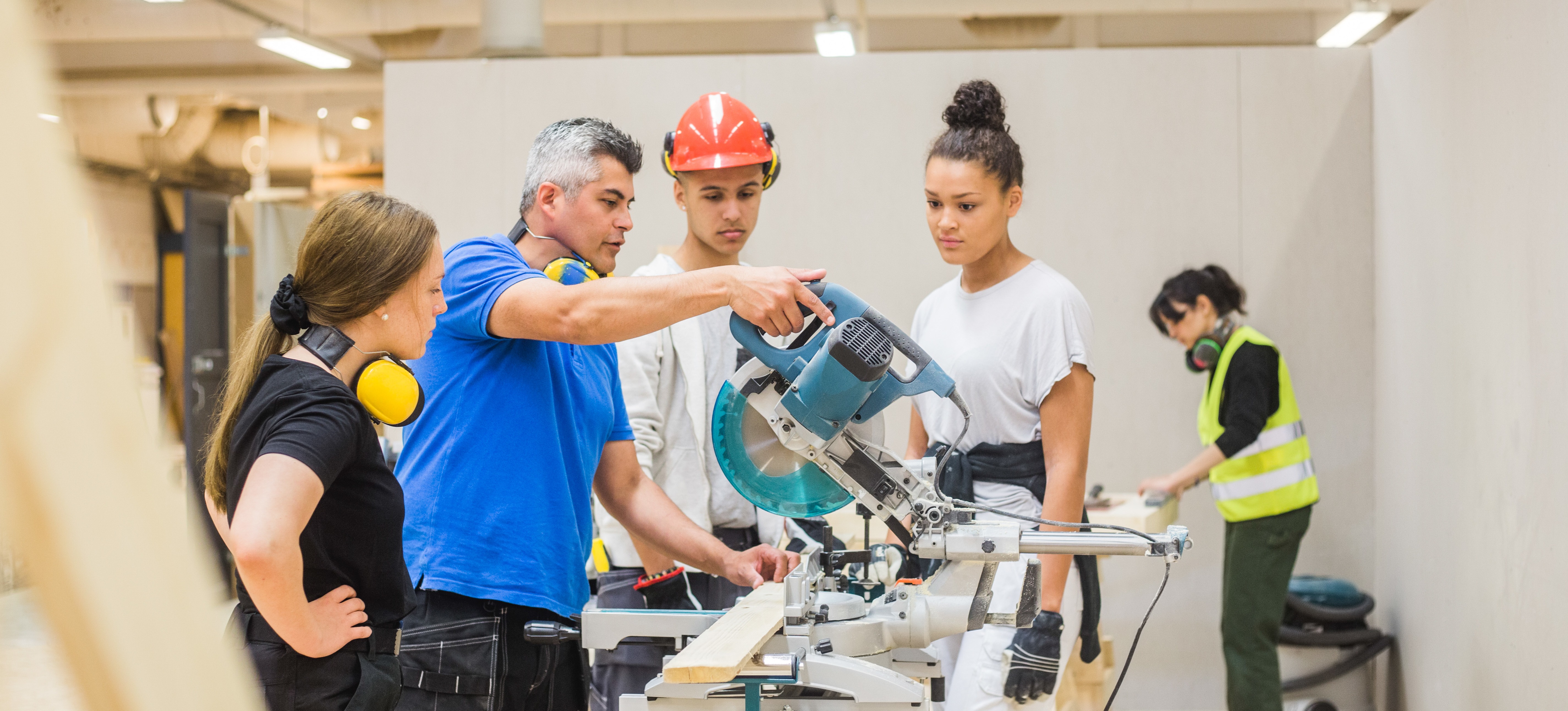 [Featured Image] A group of trade school students look on as an instructor in a construction workshop teaches them how to use a saw.
