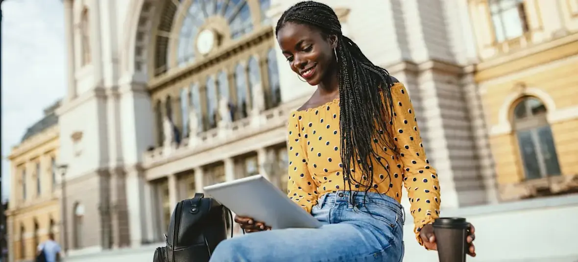 [Featured image] A young bachelor's degree student wearing a yellow polka-dot shirt sits outside drinking a coffee and looking at their tablet. 