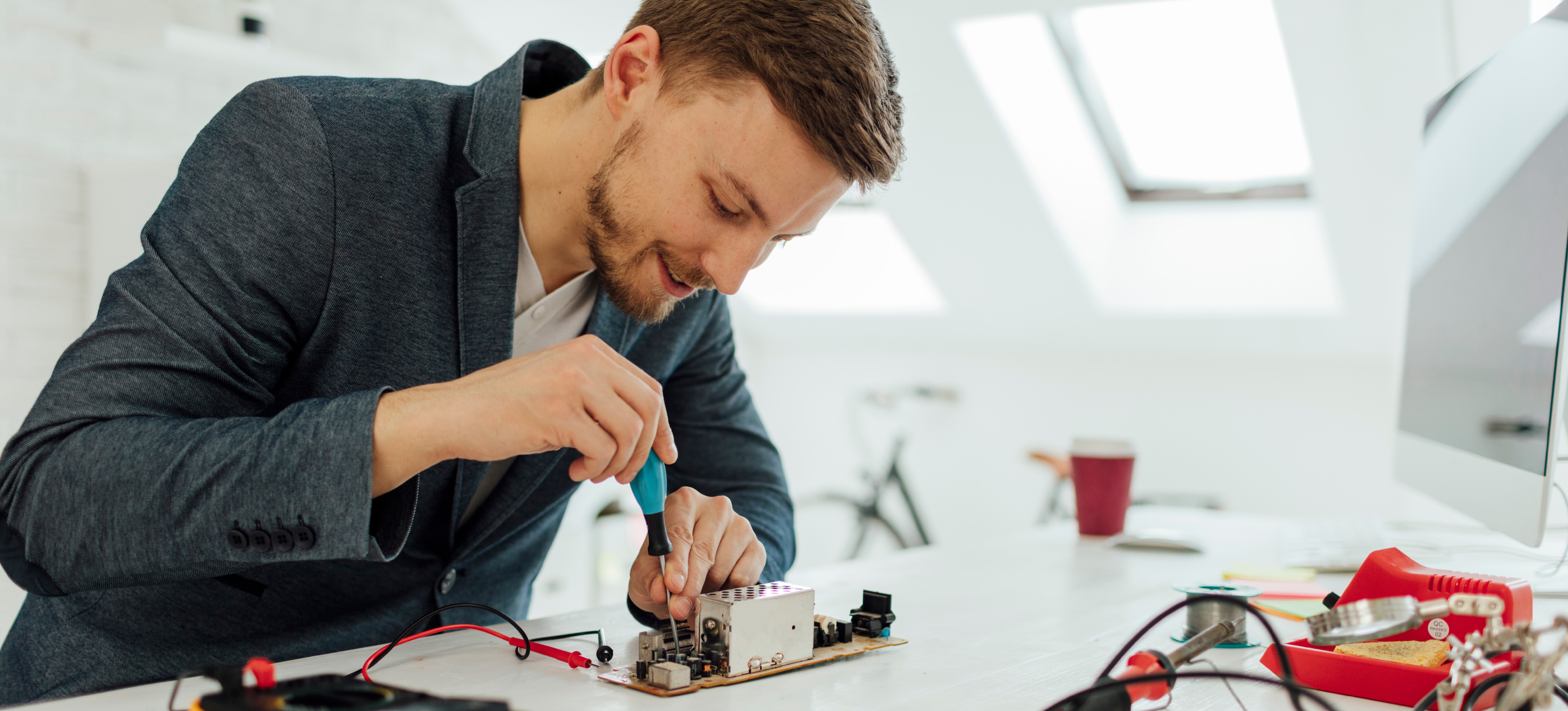 [Image en vedette] Dans son bureau, un ingénieur en matériel informatique souriant se penche sur son bureau et travaille sur un circuit imprimé à l'aide d'un tournevis.