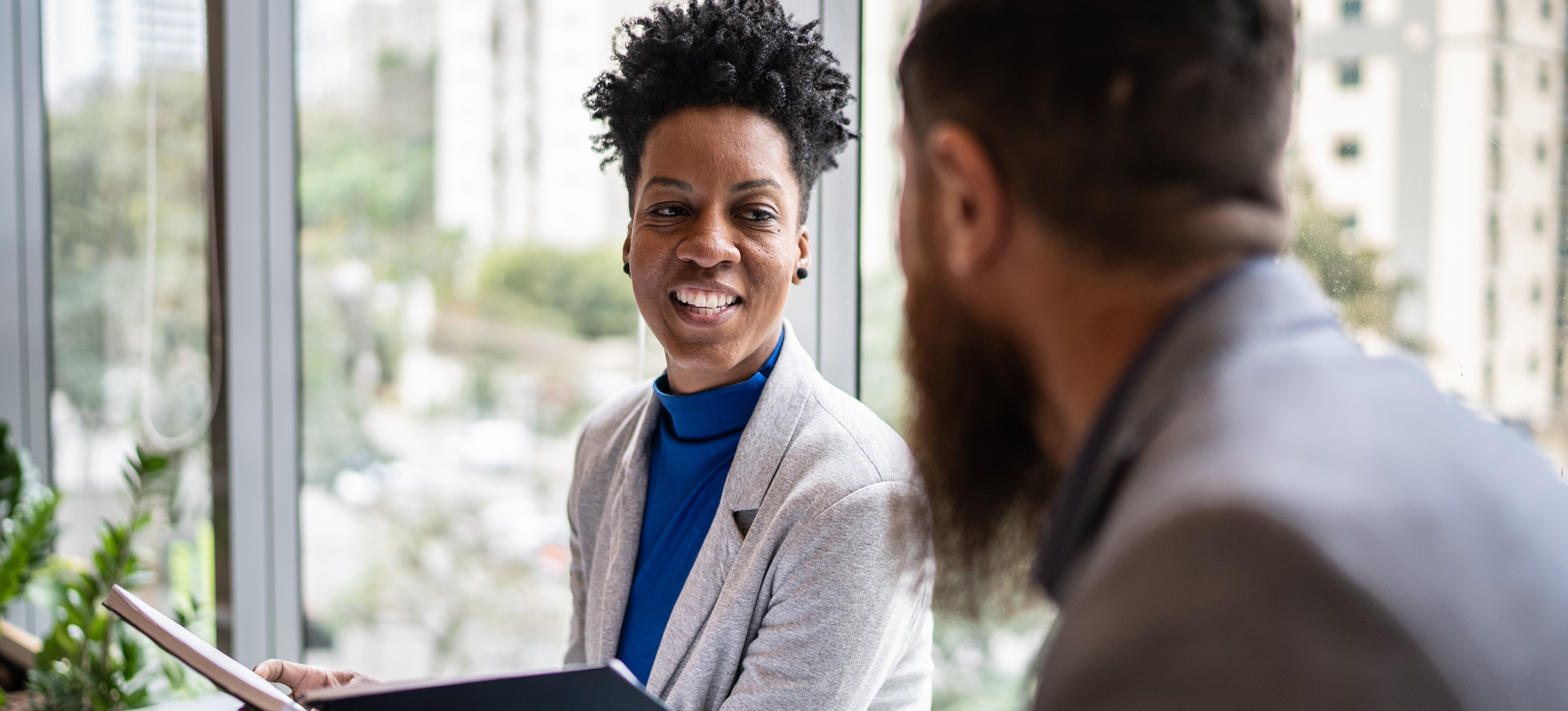 [Featured Image] A smiling HR professional holds a manual as she talks with an employee in a sunny office setting.
