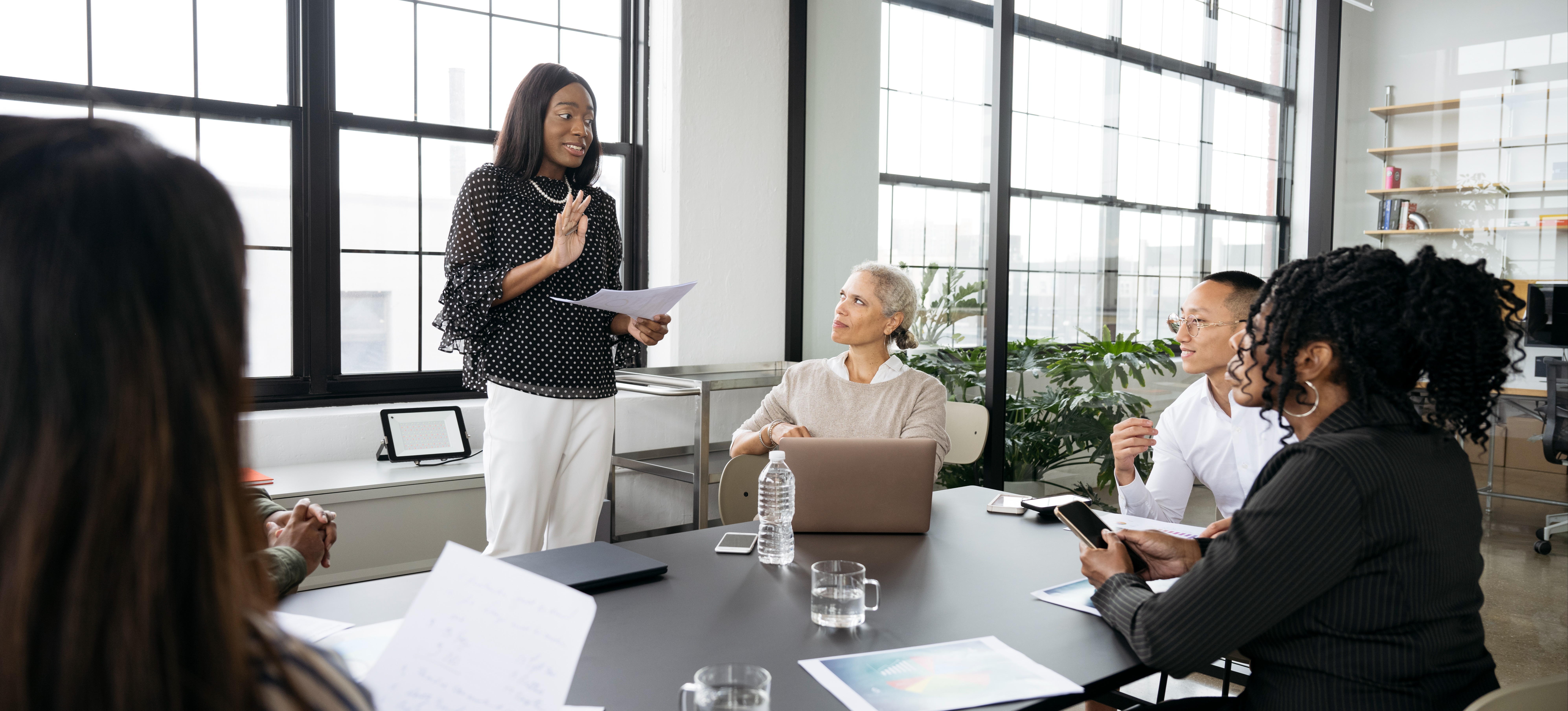 [Featured Image] A project manager stands in a sunny office space and talks to their team, who is gathered around a table, about PMI vs PMP to encourage them to seek certification as part of their employee development efforts.
