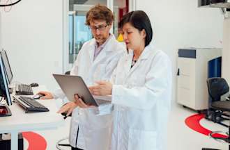 [Featured image] Two pharmacists in lab coats talk to each other while looking at a computer monitor.
