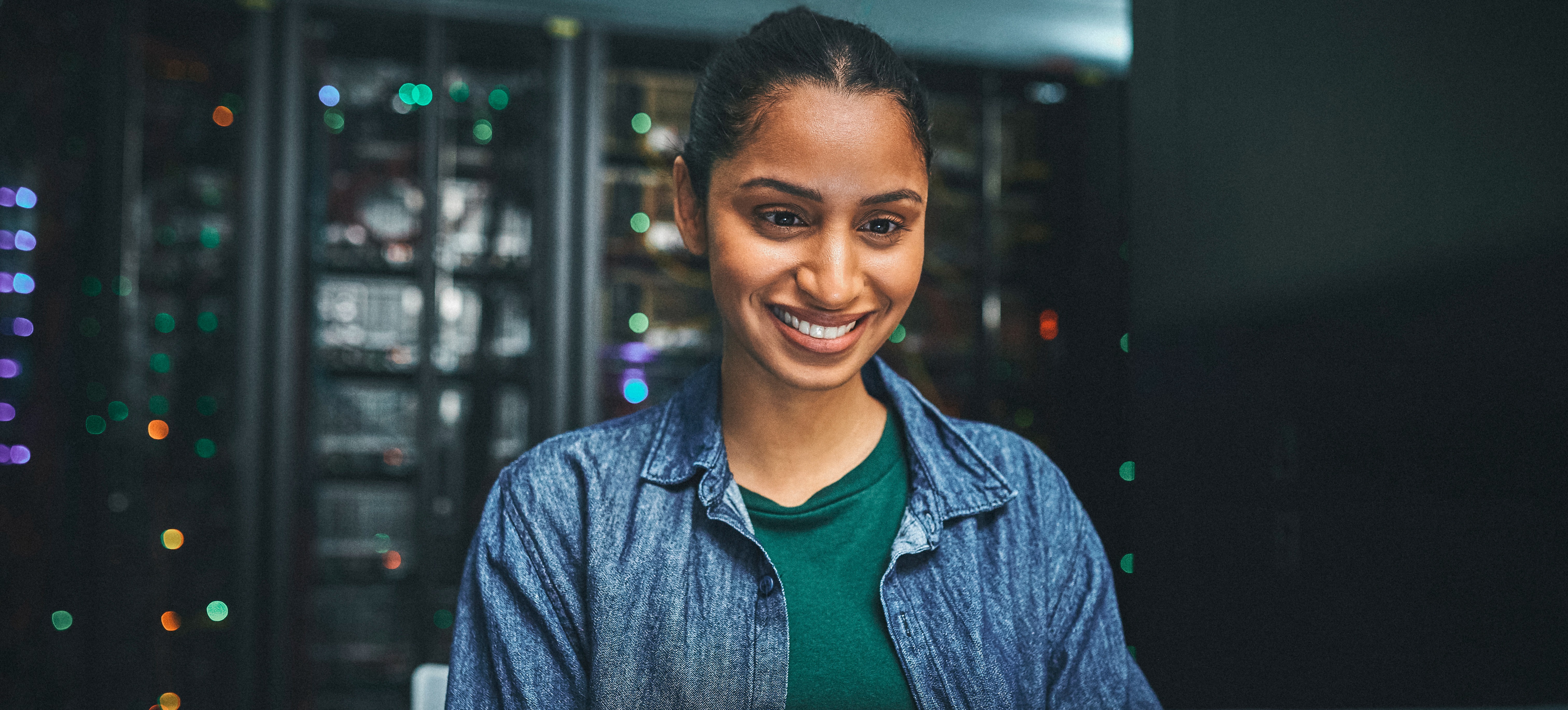 [Featured Image] A smiling cloud computing professional who has earned Google Cloud Certification works at her computer in a server room.
