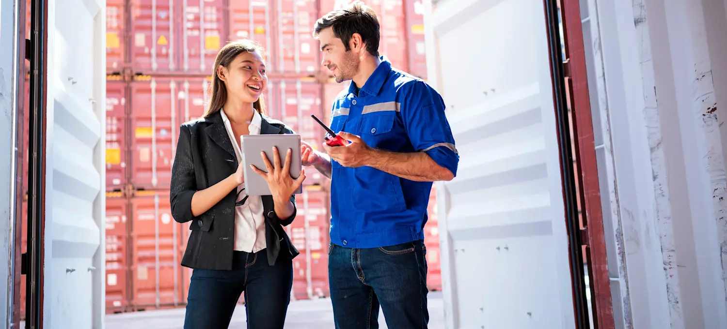 [Featured Image] Two supply chain management professionals discuss a shipment near an open freight container.

