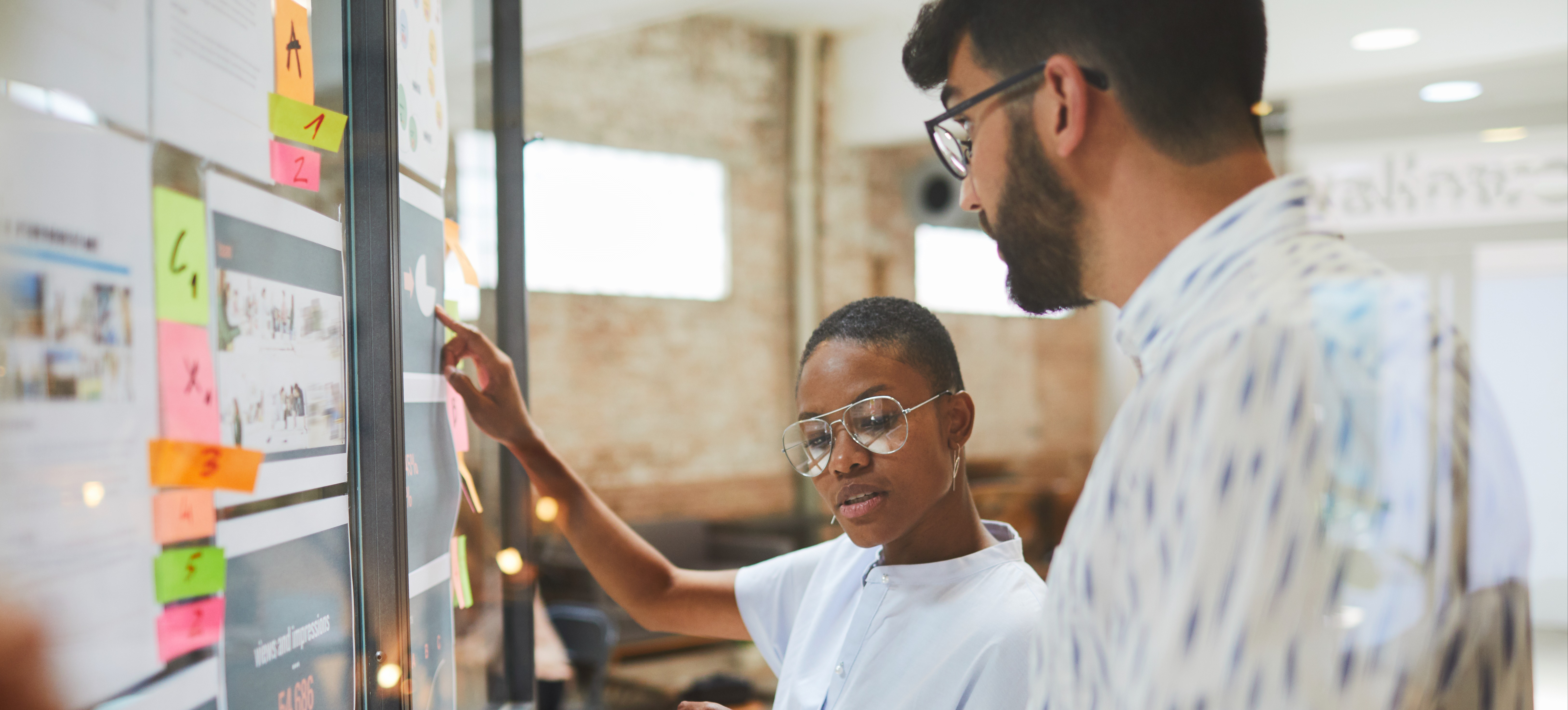 [Featured Image] Two coworkers using a display board in an office to visualize pain points. 
