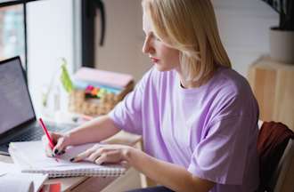 [Featured image] A student working on her entrepreneurship degree studies at a desk with a notebook, textbook, and laptop.