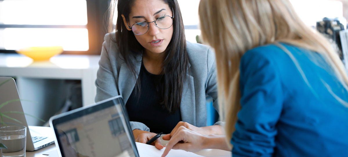 [Featured image] Two professionals sit at a table going over a competitive analysis report. Both of them have laptops in front of them.