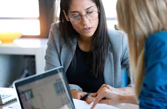 [Featured image] Two women sit at a table going over a competitive analysis report. Both of them have laptops in front of them.