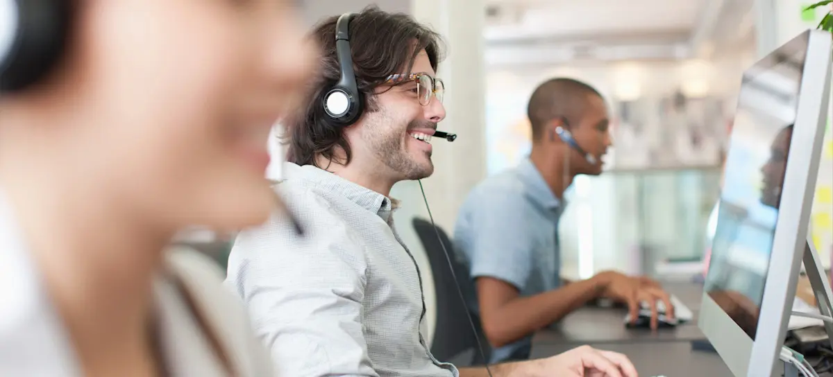 [Featured Image] Three people sit in a row, working telecommunications jobs, talking to customers on headsets and typing on their desktop computers.
