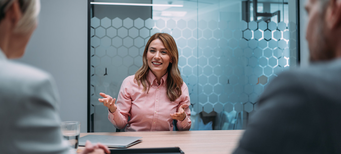[Featured Image] A person interviewee answers supply chain interview questions as she sits across a table from two interviewers in an office.
