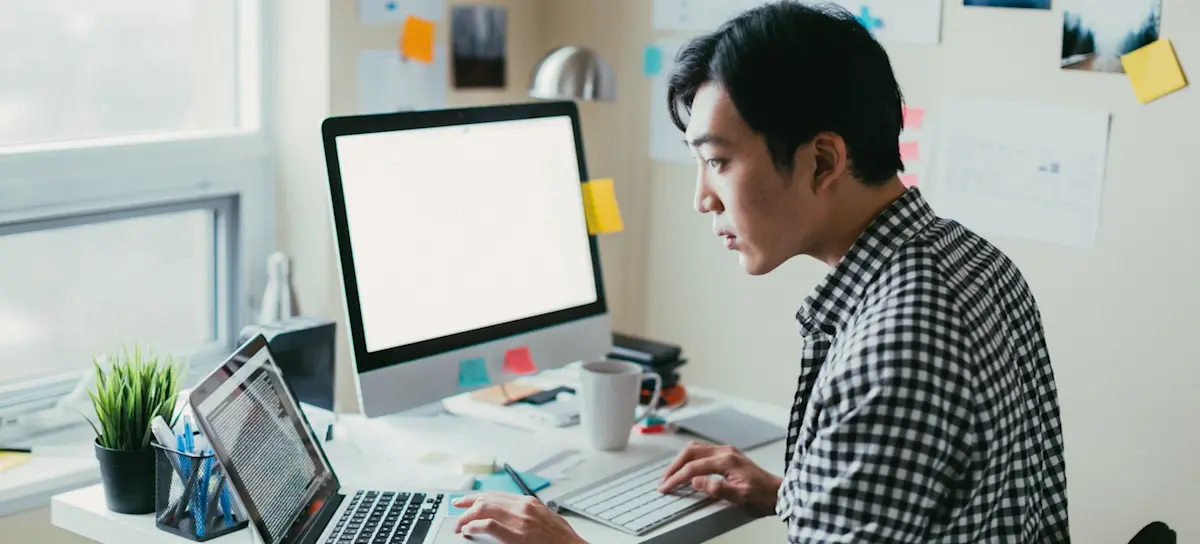 [Featured image] A CS master's degree student in a checkered shirt sits in front of a desktop computer and a laptop computer. 