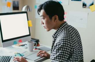 [Featured image] A young Asian man in a checkered shirt sits in front of a desktop computer and a laptop computer. 